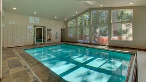 An indoor swimming pool with clear water, surrounded by large windows and a stone floor.