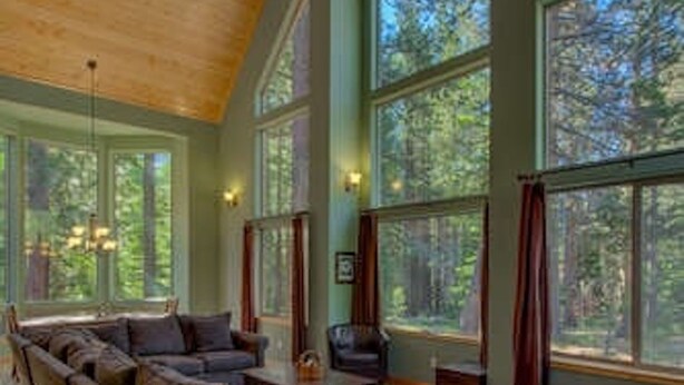 A spacious living room with a large sectional sofa, a coffee table, and a fireplace. The room has large windows offering views of the surrounding forest.
