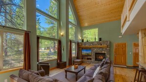 A spacious living room with a stone fireplace, large windows, and a wooden ceiling.