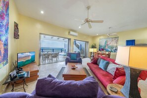 A living room with a red sofa, a coffee table, and a television on a stand. There is a balcony with a view of the ocean. - Castillito del Caribe 2 Bedrooms L1 (Isla Mujeres)