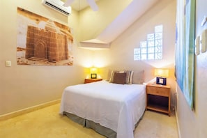 Un dormitorio con una cama, dos mesitas de noche con lámparas, una ventana y un aire acondicionado instalado en la pared.