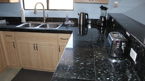 Una cocina con encimera de granito negro, pileta de acero inoxidable, alacenas de madera y cafetera.