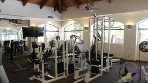 Un gimnasio con diversos equipos de ejercicio, ventanas amplias y un ventilador de techo.