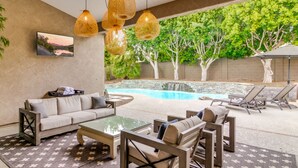 Un espace aménagé au bord de la piscine, avec un canapé, des fauteuils et une table en verre. Une piscine avec une cascade est entourée d’arbres, et un espace salon couvert est également disponible.
