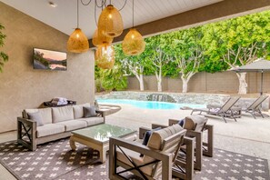 A poolside area with a sofa, chairs, and a glass table. There is a pool with a waterfall feature, surrounded by trees and a covered seating area. - SWSS 3BR Kierland Indoor-Outdoor Desert Living | Heated Pool, Hot Tub, Fire Pit & 30‑Day Minimum (Scottsdale)
