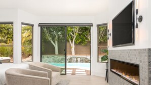 Un salon moderne doté d’une cheminée, d’un téléviseur fixé au mur et de grandes portes-fenêtres coulissantes donnant sur un jardin avec une piscine et des arbres.