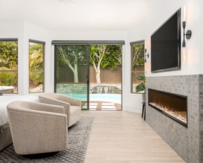A modern living room with a fireplace, a TV mounted on the wall, and large sliding glass doors leading to a backyard with a pool and trees. - SWSS 3BR Kierland Indoor-Outdoor Desert Living | Heated Pool, Hot Tub, Fire Pit & 30‑Day Minimum (Scottsdale)