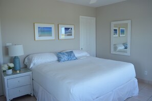A bedroom with a large bed, a nightstand with a lamp, and two framed pictures on the wall. - Beautiful 2-Bedroom, 2-Bathroom Condo: Oceanscape B1-101 (Northwest Point)