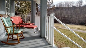 Varanda com duas cadeiras de balanço e uma mesa, com vista para uma área gramada e árvores.