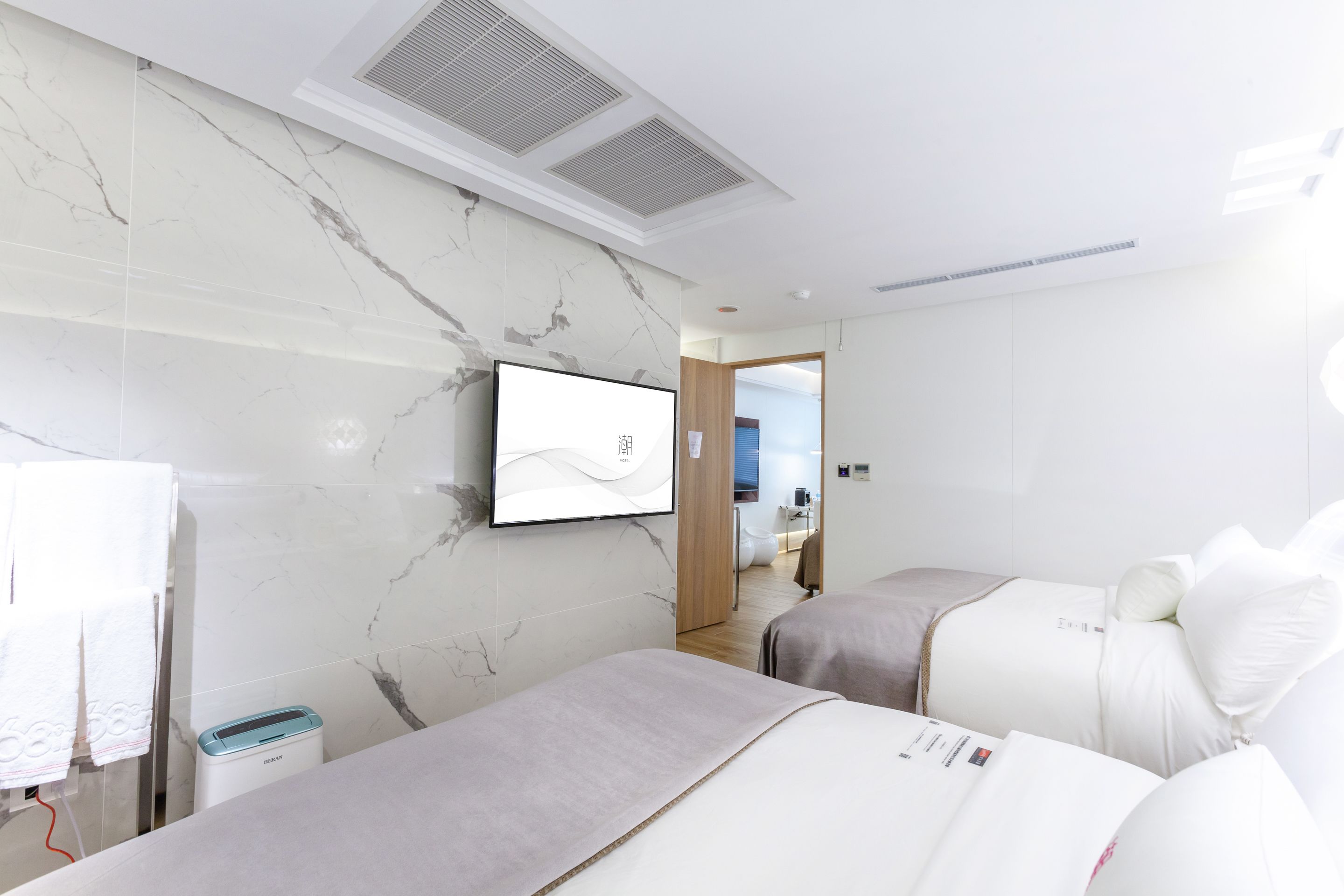 A modern hotel room with marble walls, a flat-screen TV, two beds with white bedding, a small table, and a wall-mounted heater.