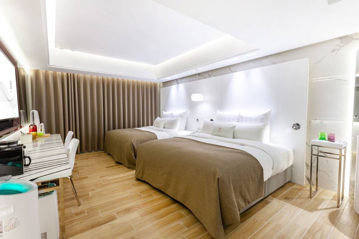 A hotel room with two beds, a desk with a chair, a small table with a mirror, and a bathroom with a shower.