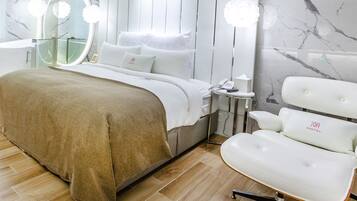 A modern bedroom with a large bed, a bedside table with a phone and tissues, a white chair, and a glass-enclosed bathtub.