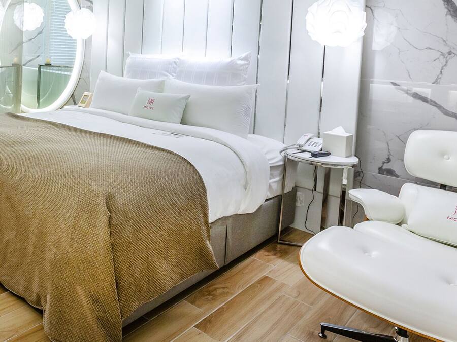 A modern bedroom with a large bed, a bedside table with a phone and tissues, a white chair, and a glass-enclosed bathtub.