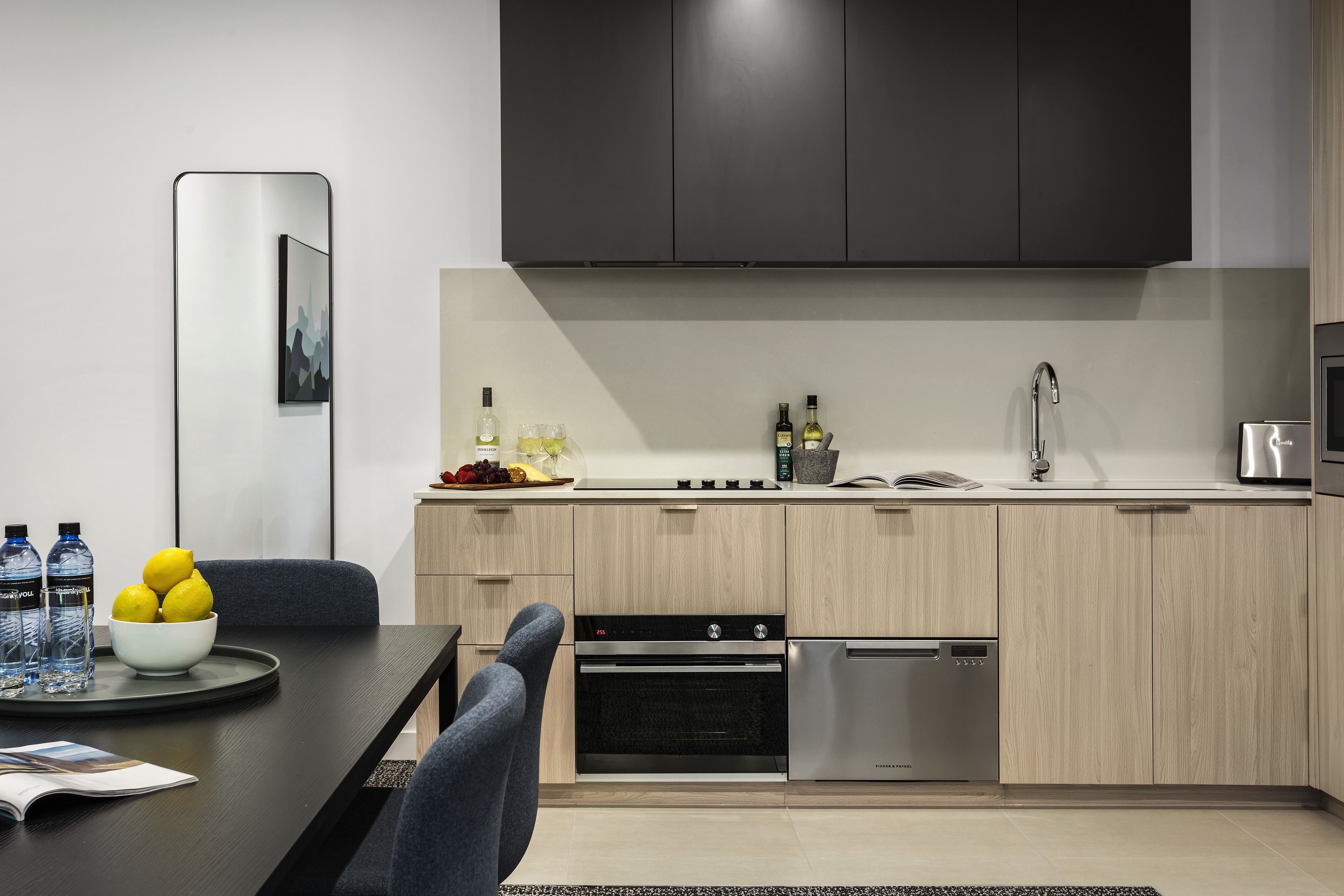 A modern kitchen with light wood cabinets, a black countertop, stainless steel appliances, and a dining area with a table and chairs.