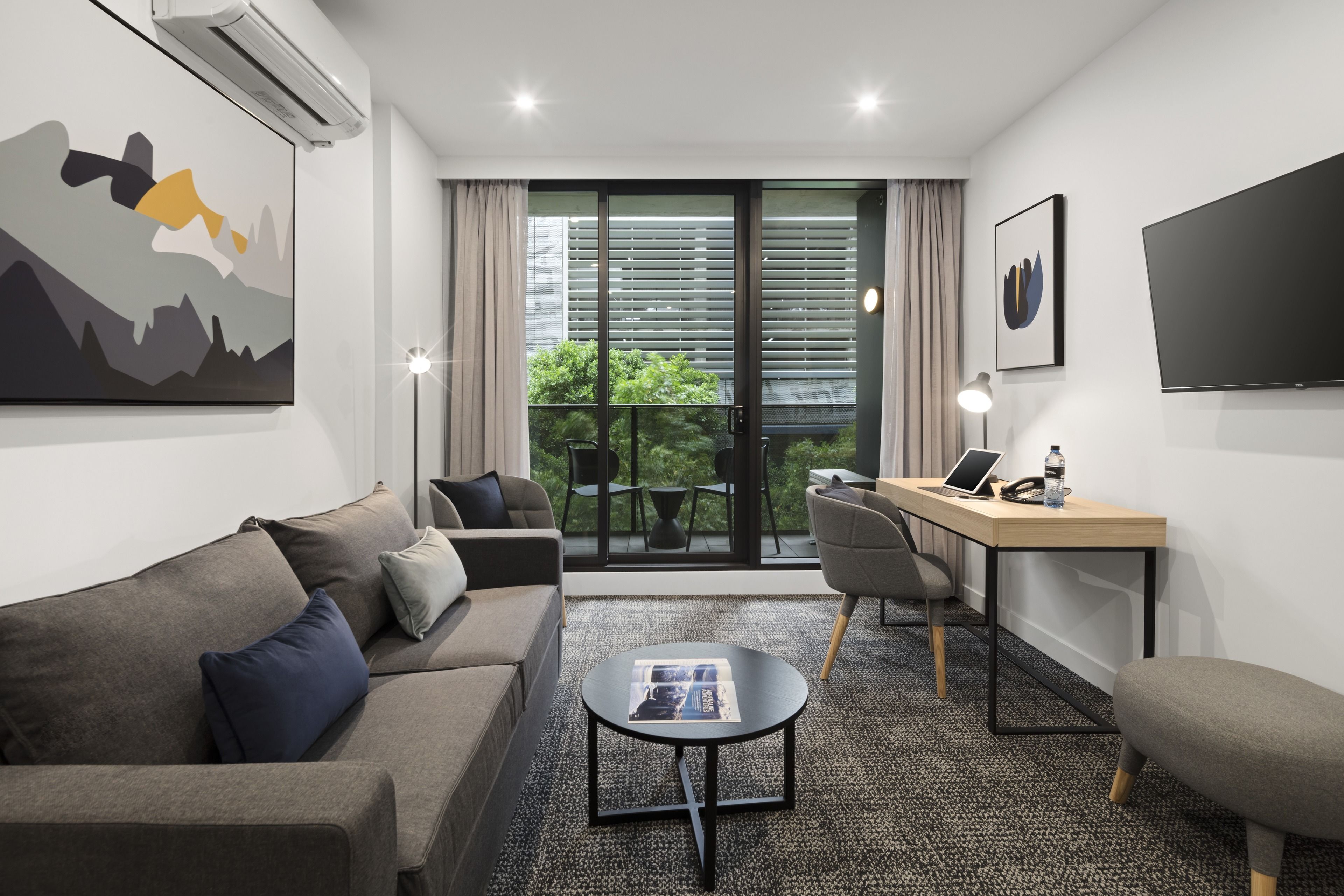 A modern hotel room with a sofa, a small round table, a desk with a laptop, and a large window with blinds.