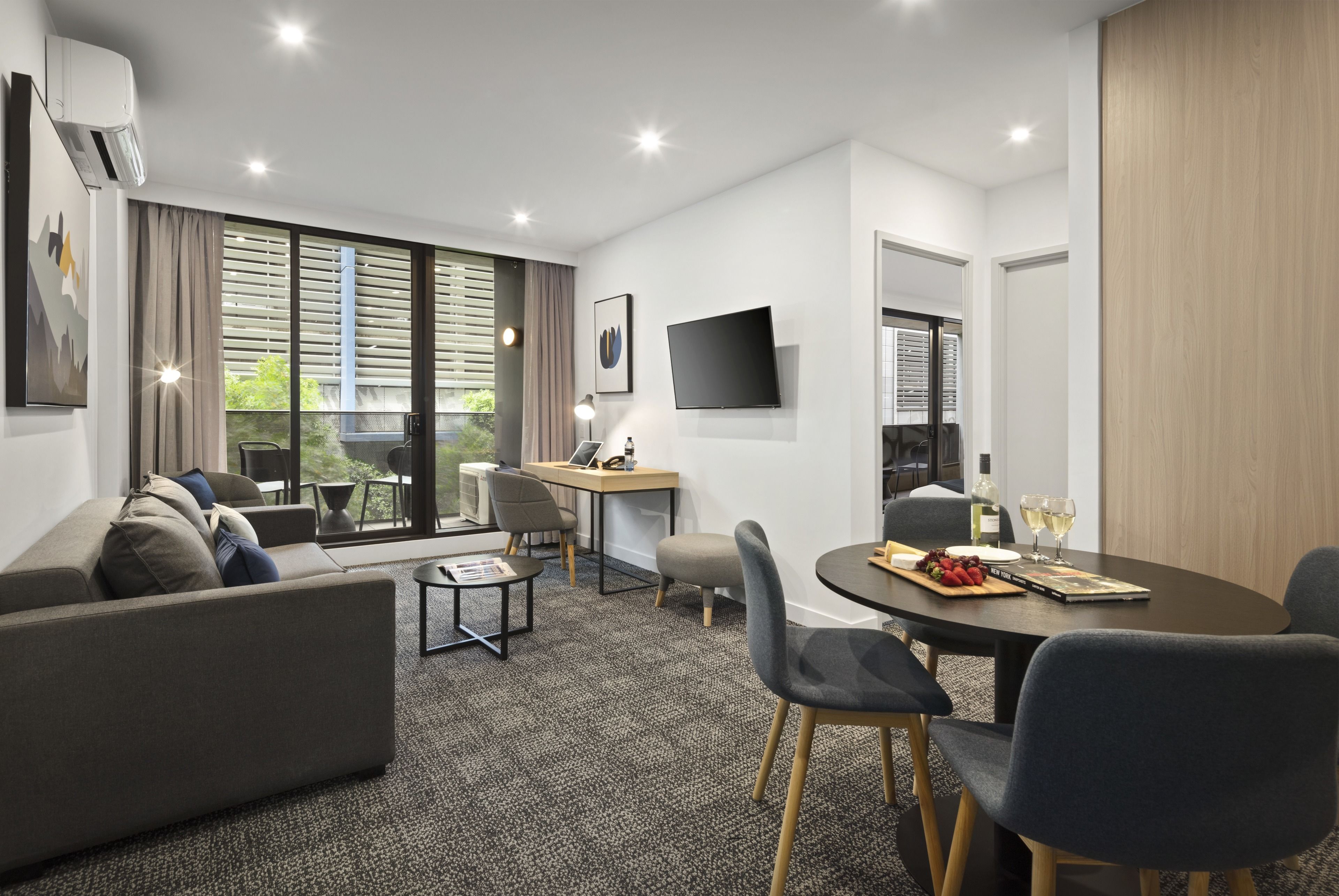 A modern living room with a sofa, a dining table with chairs, a flat-screen TV mounted on the wall, and large windows with blinds.