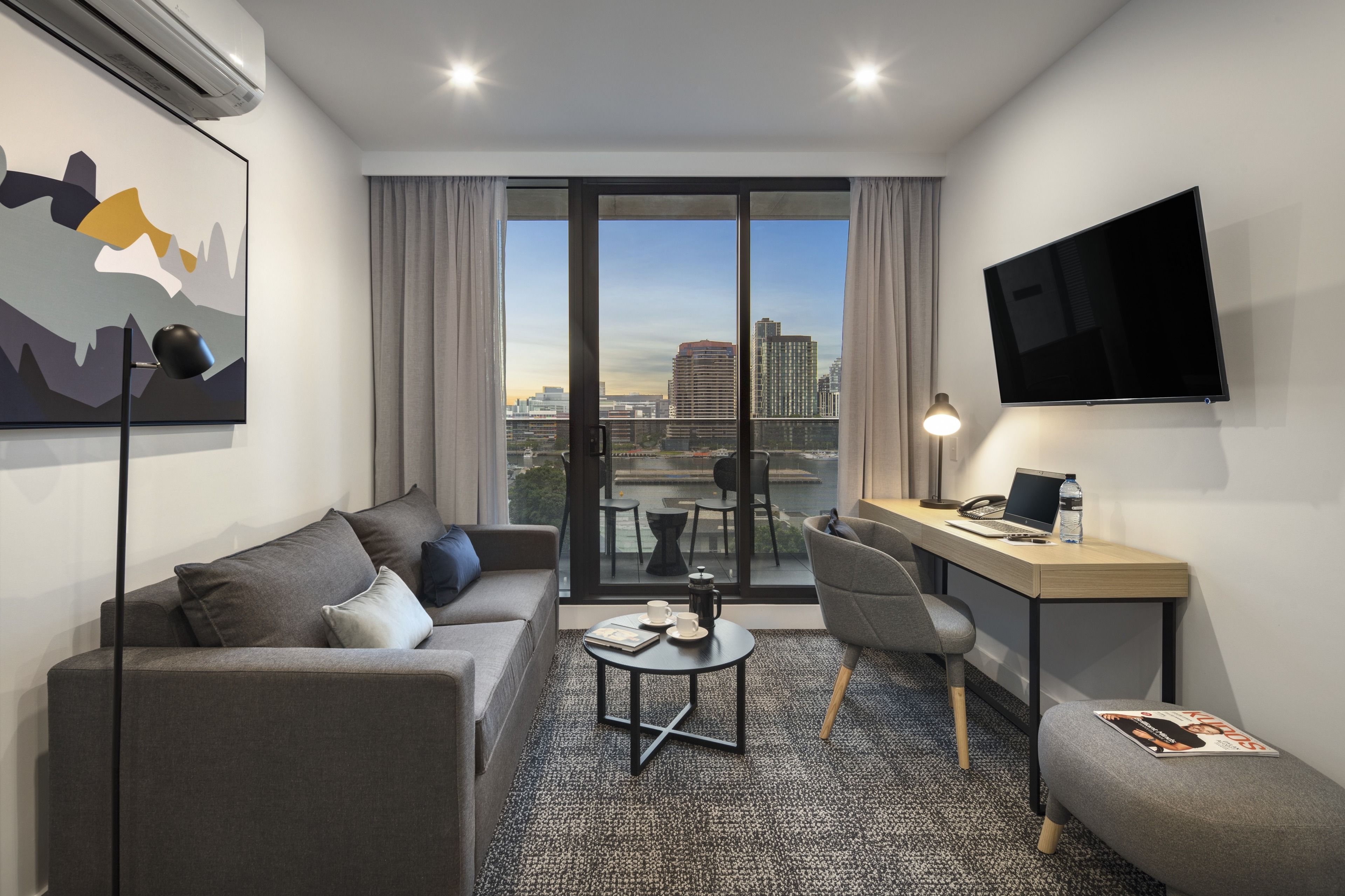 Una habitación de hotel moderna con sofá, una pequeña mesa redonda, escritorio, silla, televisor y un ventanal con vistas a la ciudad.