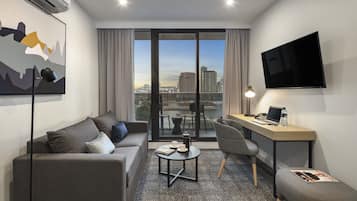 Una habitación de hotel moderna con sofá, una pequeña mesa redonda, escritorio, silla, televisor y un ventanal con vistas a la ciudad.