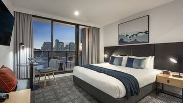 Ett modernt hotellrum med en stor säng, ett skrivbord med en stol, ett litet bord och utsikt över stadens silhuett genom stora fönster som sträcker sig från golv till tak.