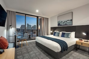 A modern hotel room with a large bed, a desk with a chair, a small table, and a view of the cityscape. - Quest NewQuay (Docklands)