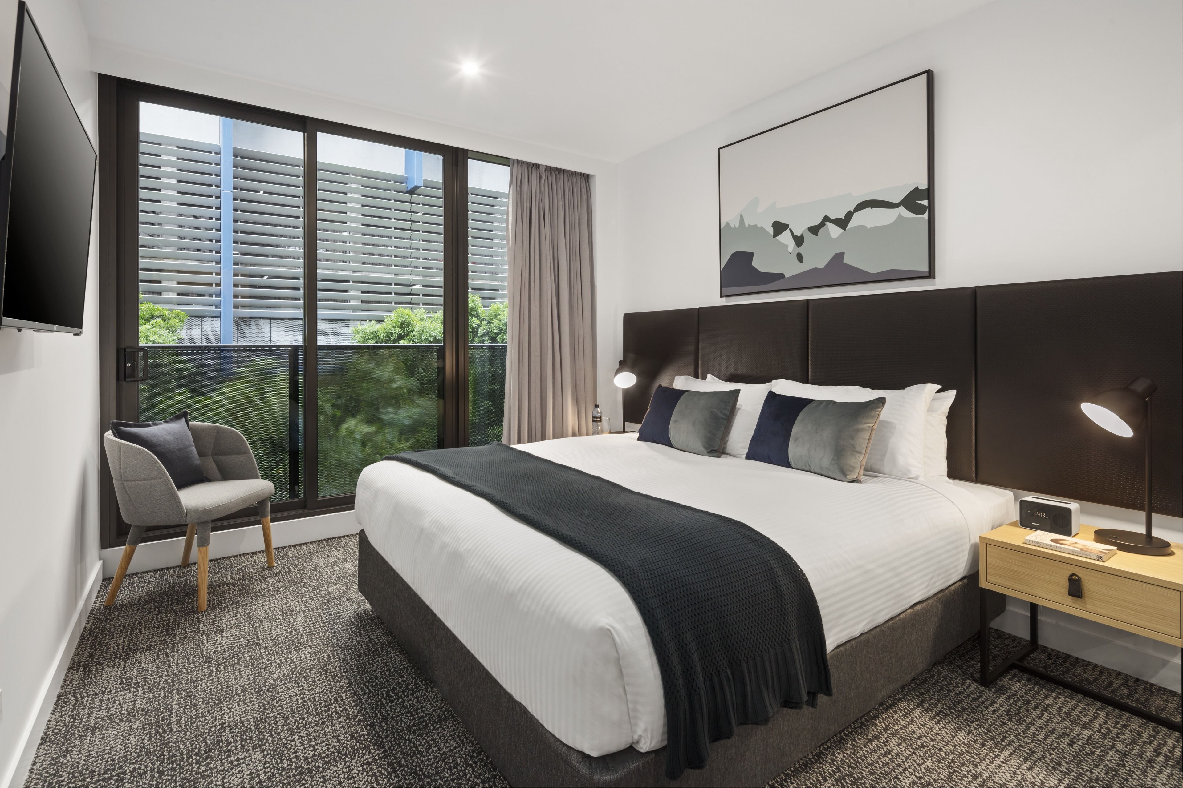 A modern hotel room with a large bed, a chair, a nightstand with a lamp, and a view of the city through large windows.