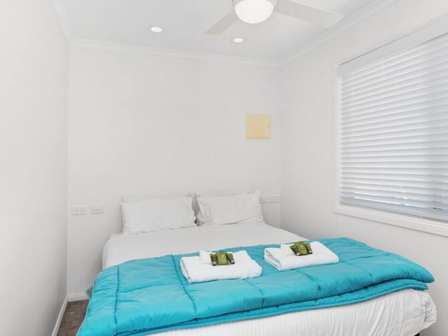 Quarto de hotel com cama, ventilador de teto, janela com persianas e porta com puxador.