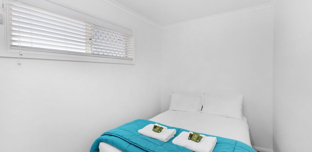 A white room with a bed, a window with blinds, and a blue blanket on the bed.