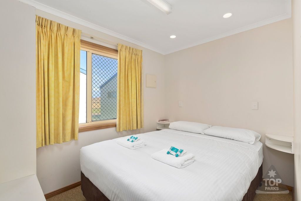 A hotel room with a bed, two towels, a nightstand, a window with yellow curtains, and a view of a house.