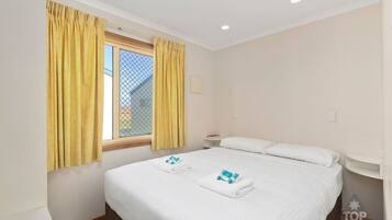 A hotel room with a bed, two towels, a nightstand, a window with yellow curtains, and a view of a house.