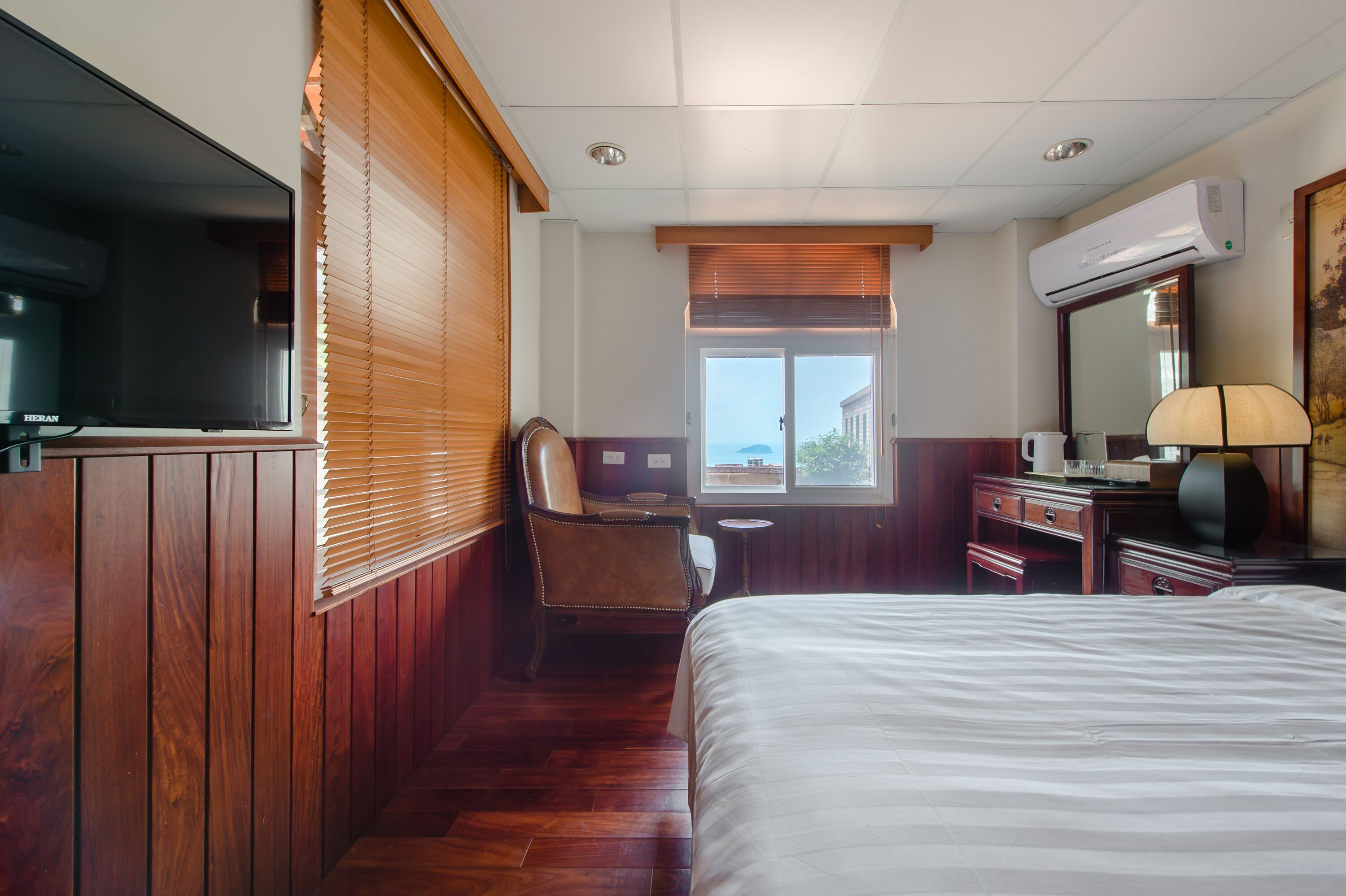 A hotel room with a large bed, a desk with a chair, a television, an air conditioning unit, and a window with a view.