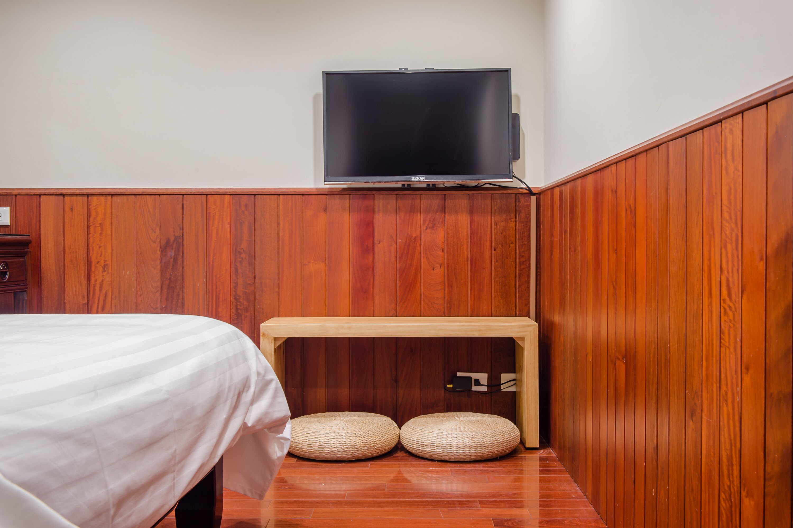 A small room with a bed, a television mounted on the wall, and a wooden bench with two cushions.