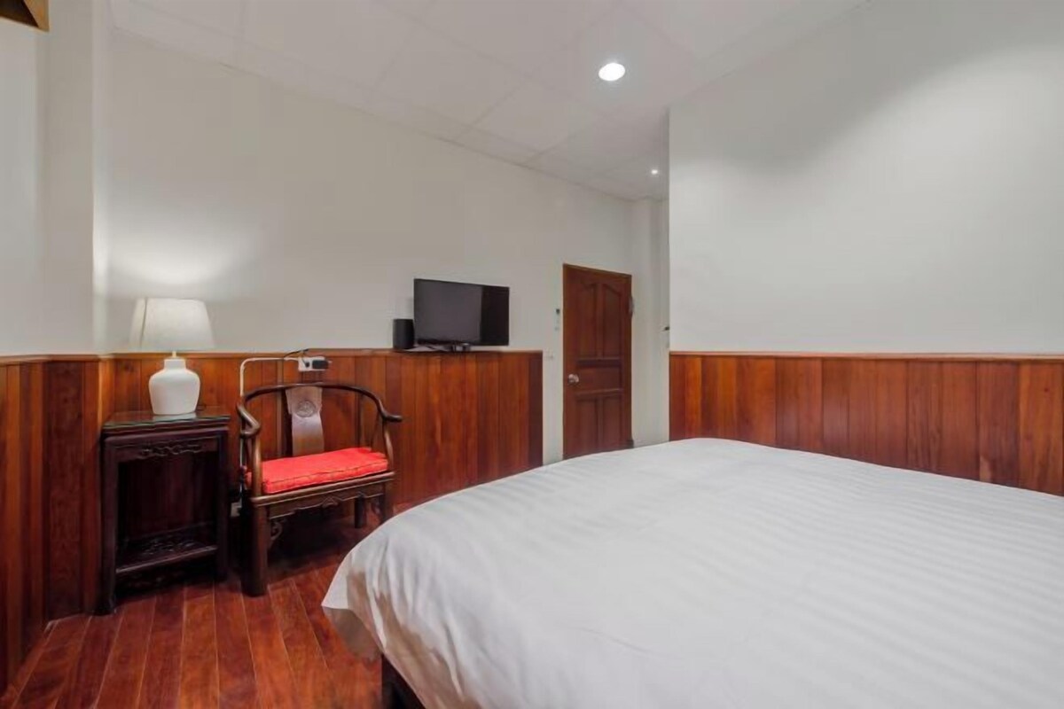 A hotel room with a wooden paneled wall, a bed with white bedding, a small bedside table with a lamp, a chair, and a television on the wall.