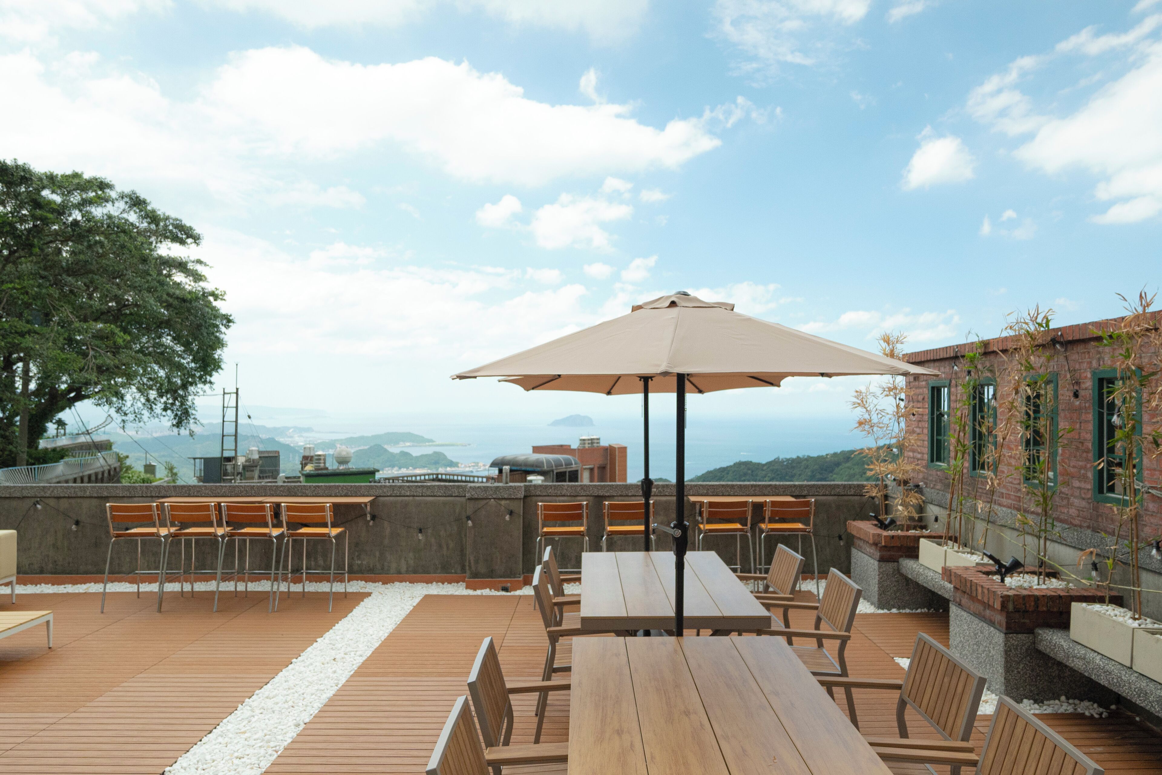 Un patio al aire libre con mesas y sillas de madera, un paraguas color beige y vistas del paisaje urbano.