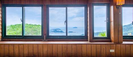 A traditional Japanese room with tatami flooring, a wooden ceiling, and large windows offering a view of greenery and the sea.