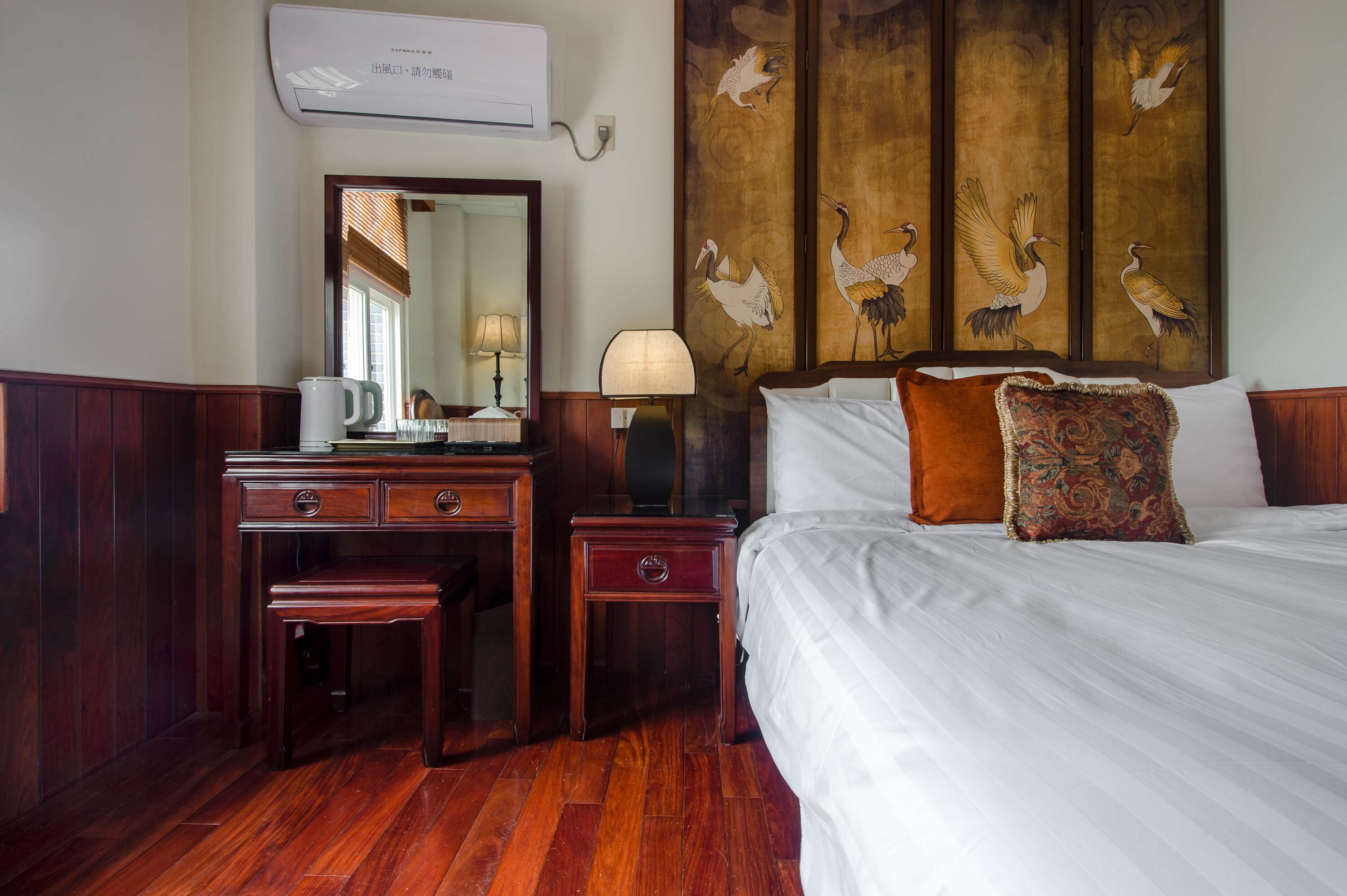 Habitación de hotel con un escritorio de madera, un espejo, una lámpara, una cama con ropa blanca y dos almohadas, y una pared con un cuadro de pájaros.