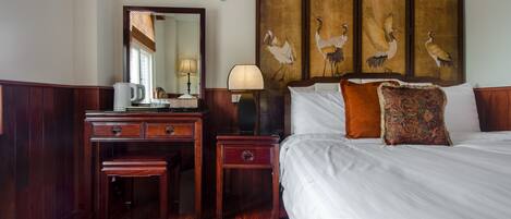 Habitación de hotel con un escritorio de madera, un espejo, una lámpara, una cama con ropa blanca y dos almohadas, y una pared con un cuadro de pájaros.