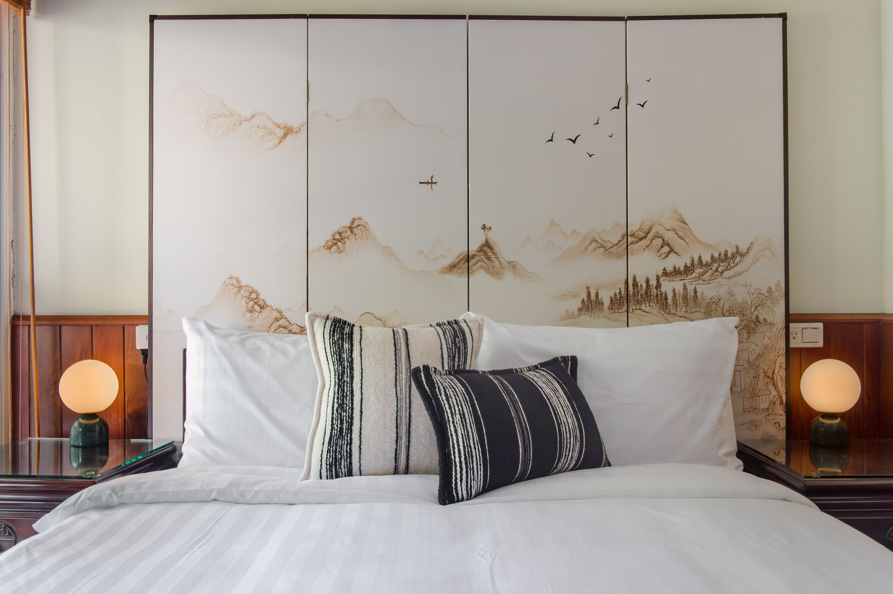 A hotel room with a bed, two pillows, a nightstand with a lamp, and a wall with a traditional artwork.