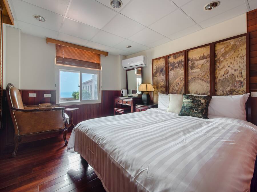 Habitación de hotel con una cama grande, una silla de madera, un escritorio con lámpara y una ventana con persianas.
