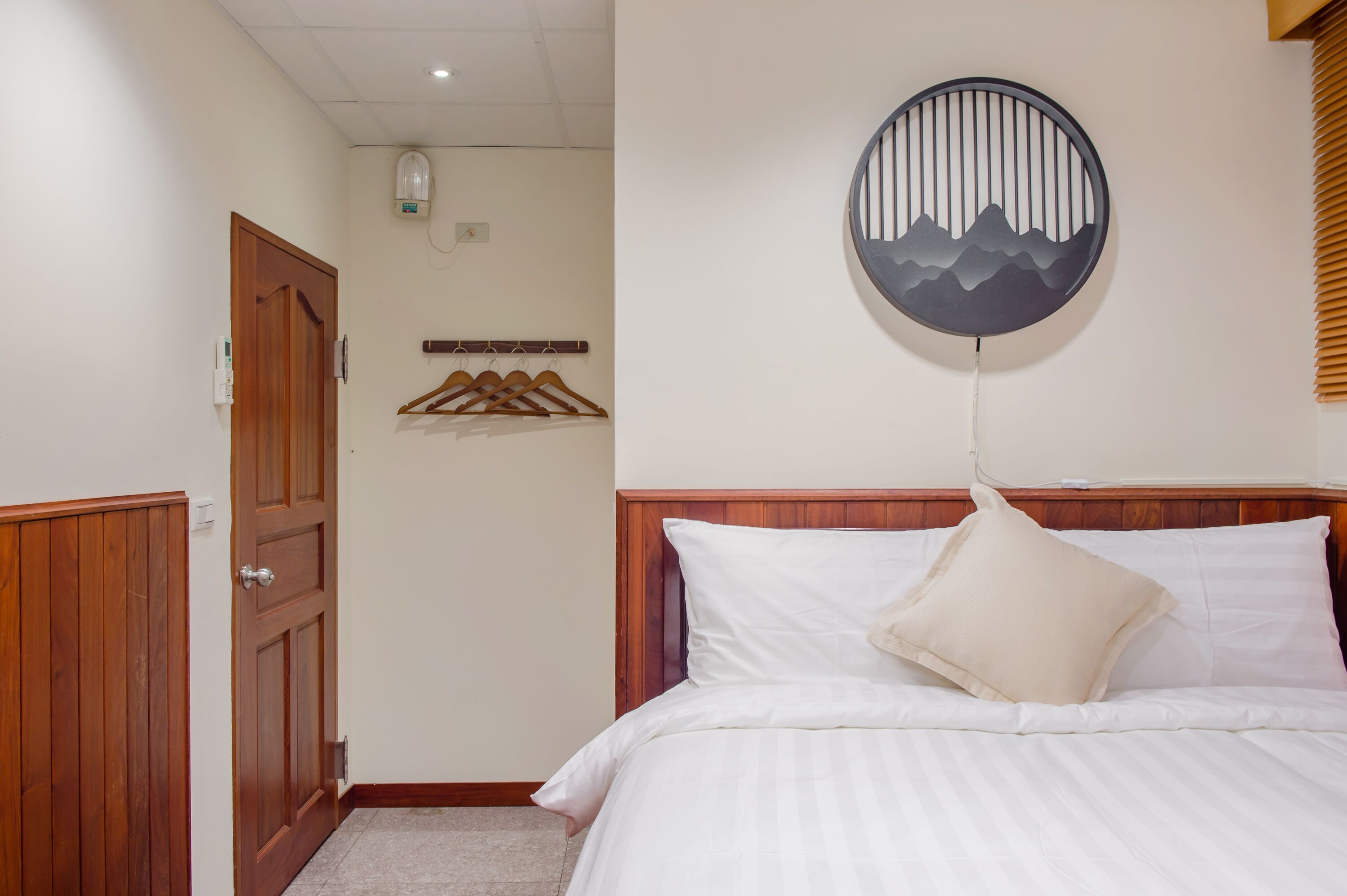 Une chambre avec une porte en bois, un lit avec une literie blanche, un luminaire fixé au mur et une œuvre d’art murale de forme circulaire.