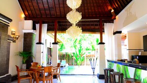 A spacious indoor area with a dining table and chairs, a bar area with stools, and a view of a pool and greenery outside. - Villa Kubu Tanjung Sanur Bali (Denpasar)