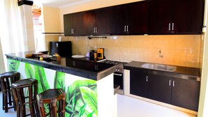 A modern kitchen with dark cabinets, a black countertop, and a green leaf-patterned backsplash. There are three wooden stools and a built-in bar area. - Villa Kubu Tanjung Sanur Bali (Denpasar)