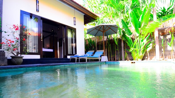 A swimming pool with clear water, surrounded by lush greenery and a modern building with a large window and a door. - Villa Kubu Tanjung Sanur Bali (Denpasar)