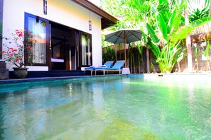 A swimming pool with clear water, surrounded by lush greenery and a modern building with a large window and a door. - Villa Kubu Tanjung Sanur Bali (Denpasar)