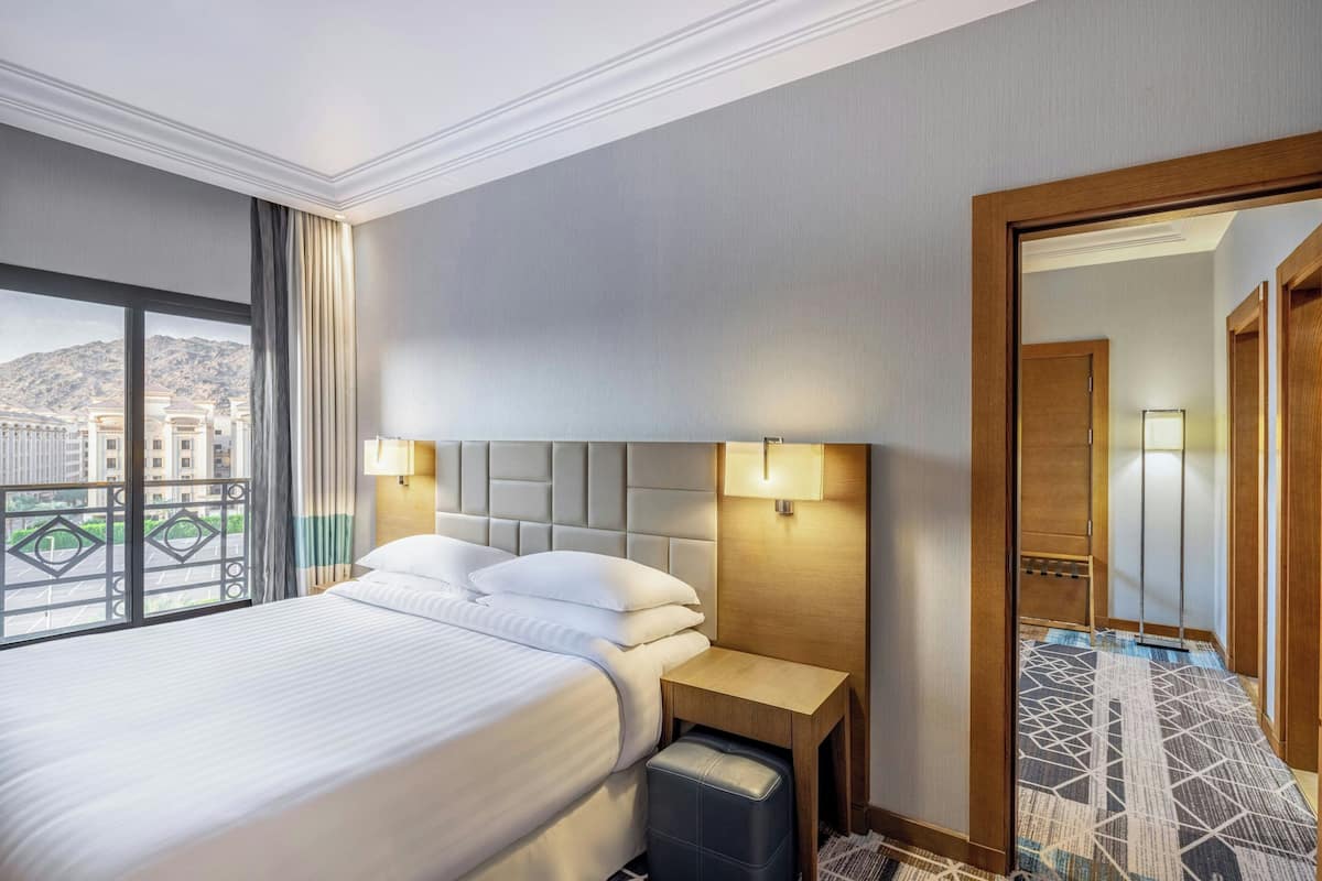 Una habitación de hotel con una cama grande, dos lámparas de noche, una mesita de noche de madera y vistas a la ciudad a través de un ventanal amplio.