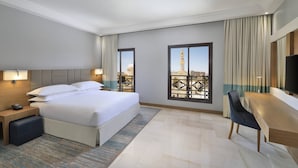 Habitación de hotel con una cama grande, un escritorio y vistas a un paisaje urbano a través de las ventanas.