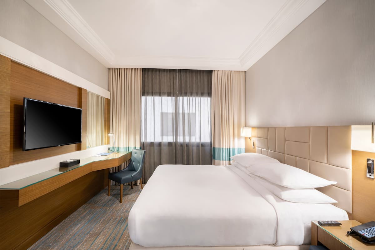 Una habitación de hotel con una cama grande, un escritorio con televisor de pantalla plana, una silla y una mesita de noche con lámpara.