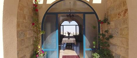 An arched doorway leading to a room with a table and a bench, surrounded by stone walls and adorned with flowers.