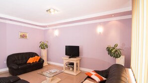 Un salon avec un canapé en cuir noir, une table basse en verre, un téléviseur posé sur un meuble en bois et deux plantes en pot.