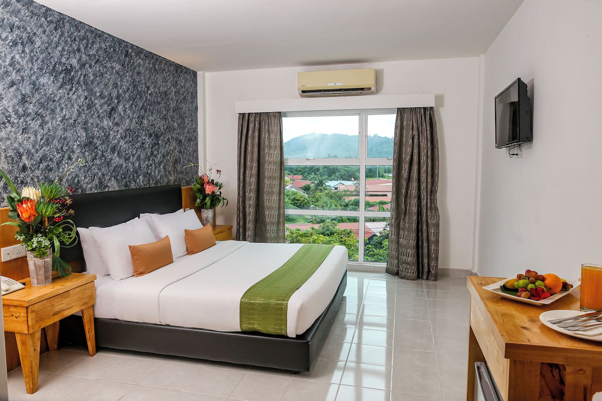 A hotel room with a large bed, a wooden nightstand, a desk with a plate of fruit and a glass of juice, a wall-mounted TV, and a window with curtains.