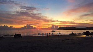Cena de praia ao pôr do sol com silhuetas de pessoas e barcos.